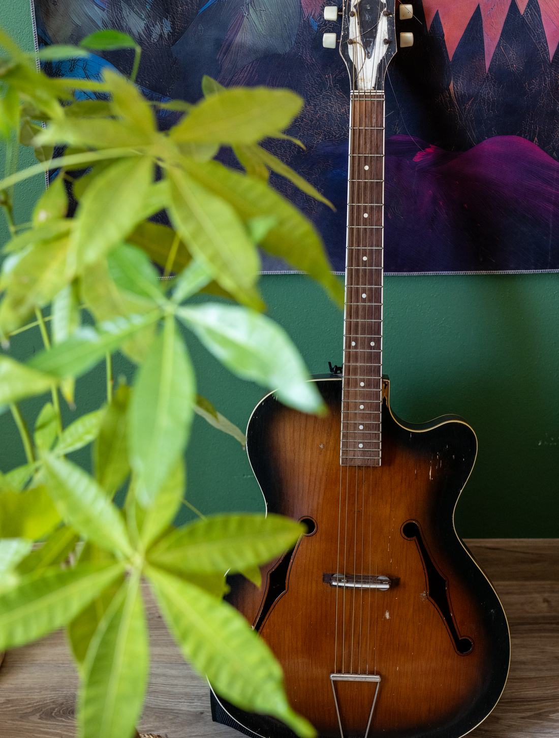 Gitara oparta o zieloną ścianę zielonej strefy, przed nią liście rośliny doniczkowej, w tle malowana tkanina