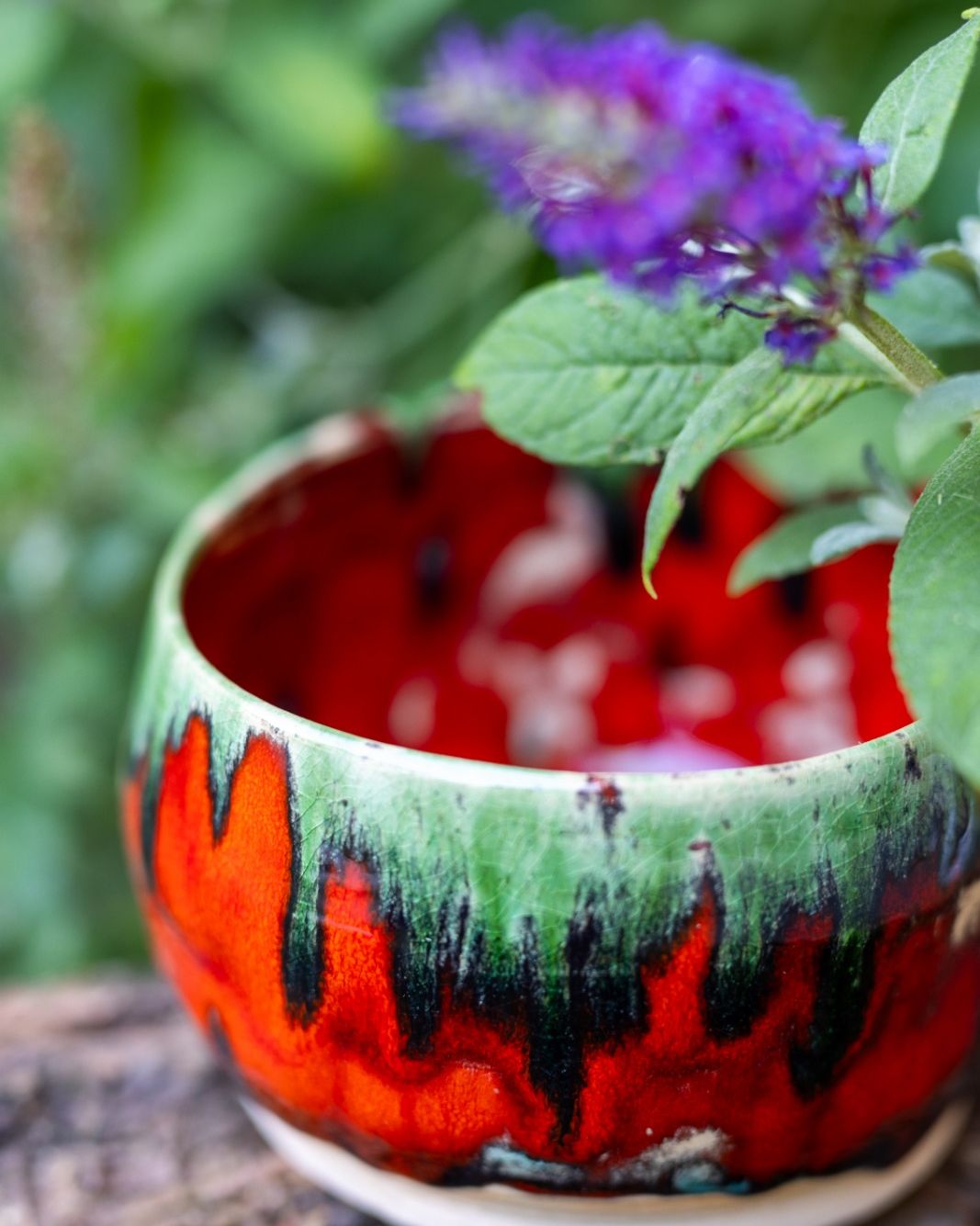 Ceramiczna misa z czerwono-zielonym szkliwem stojąca na korze, nad nią gałązka z fioletowym kwiatem